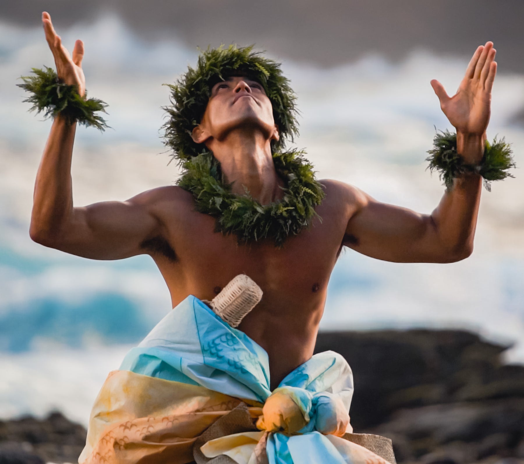 Man in traditional attire with leis and a fish hook on a beach